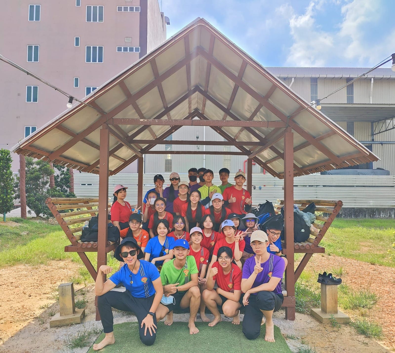 A large group of young people posing together under an outdoor wooden gazebo with buildings in the background.